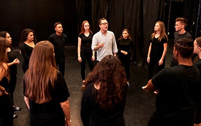 A group of performers in black attire gathers in a circle, engaged in a discussion led by a man in a light shirt. The setting is a rehearsal space, emphasizing collaboration in the arts.