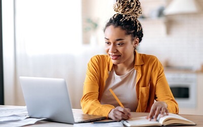 A young woman with braided hair smiles while using a laptop, taking notes in a notebook. She appears focused in a cozy workspace, reflecting a productive environment.