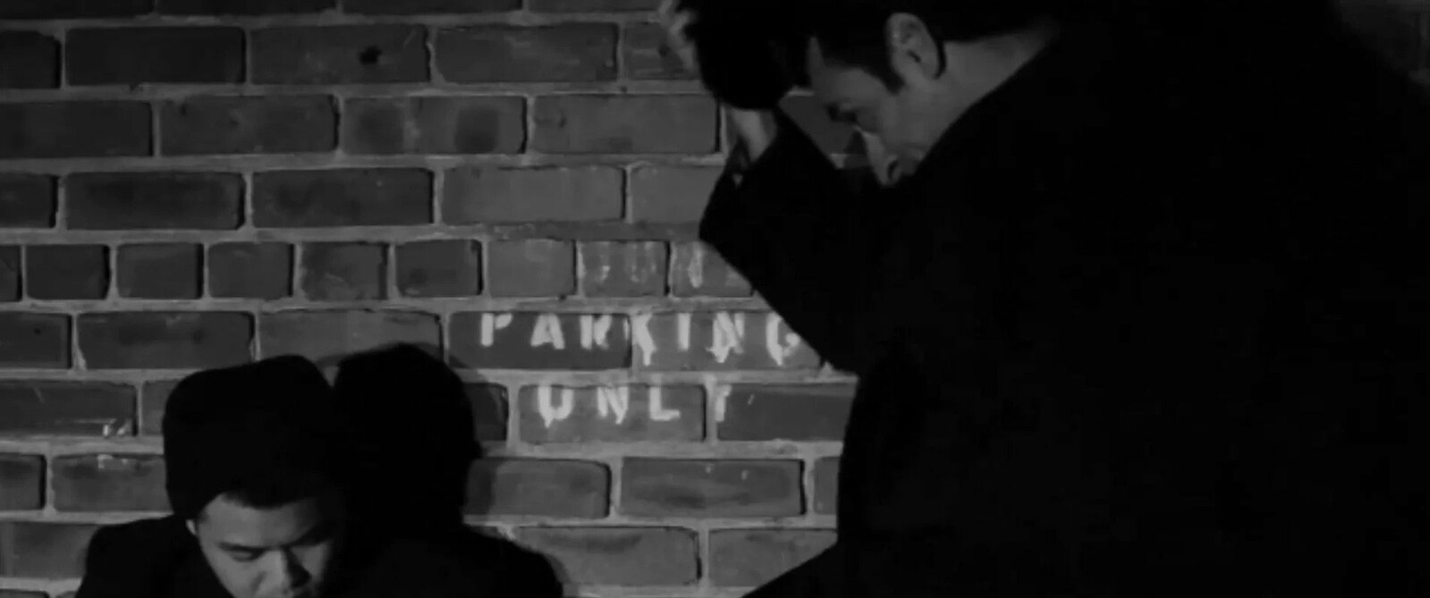 Two men in dark clothing lean against a brick wall, one appearing distressed while the other is partially obscured. The wall bears a "Parking Only" sign.