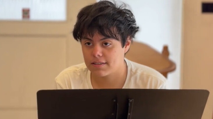 A young person with short, dark hair sits attentively at a music stand in a bright room, engaged in a rehearsal or practice session.