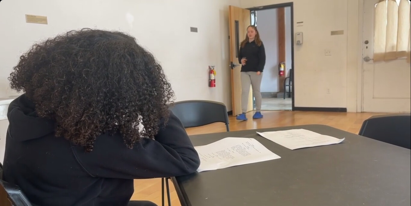 A person with curly hair sits at a table with papers, while another individual enters the room, preparing for a discussion or presentation.