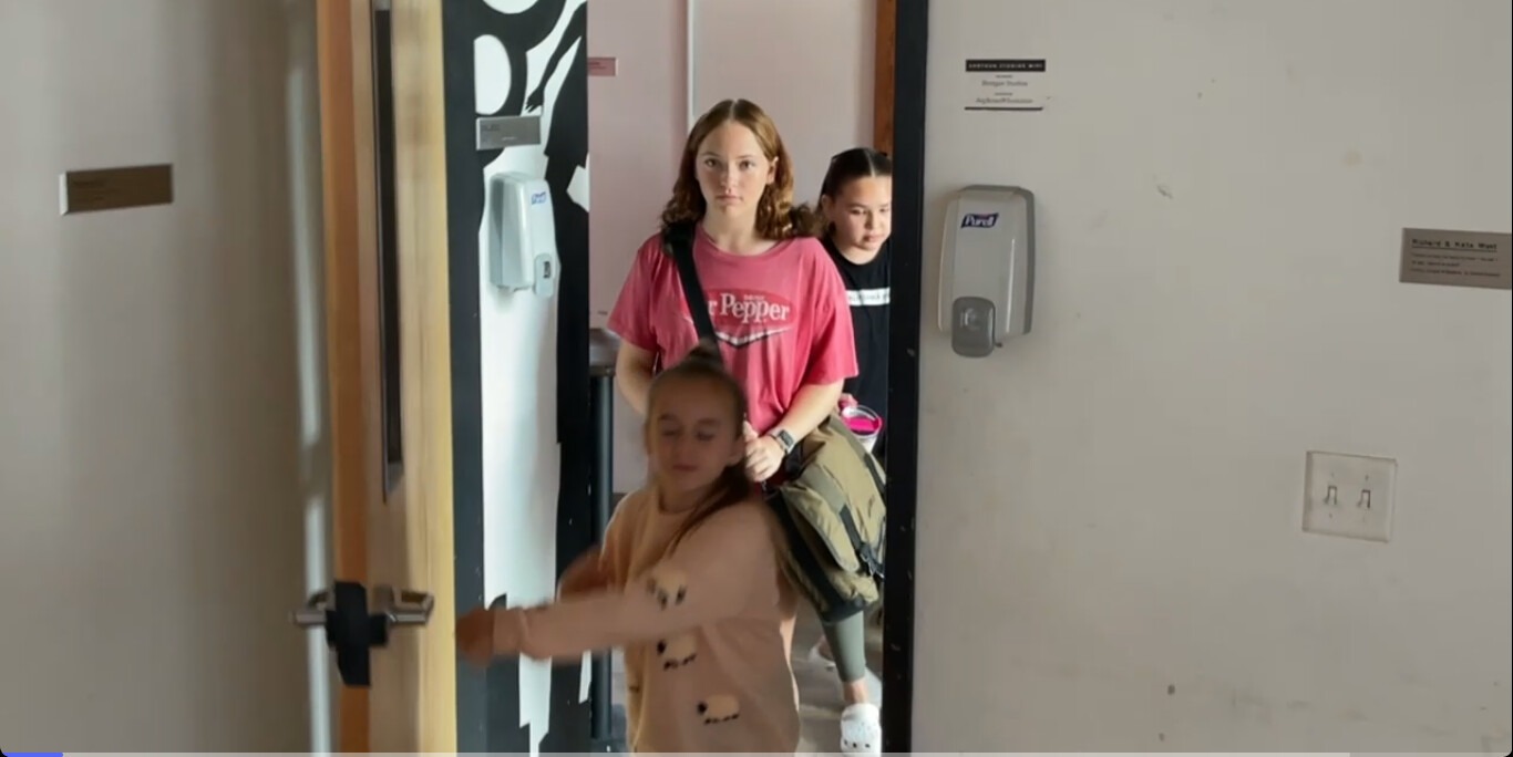 Three children enter a room, with one girl at the forefront wearing a pink shirt. The setting is a hallway with a hand sanitizer dispenser visible.