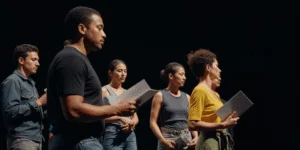 Actors perform a reading on stage, holding scripts under dim lighting, emphasizing their expressions and the focus of the rehearsal.