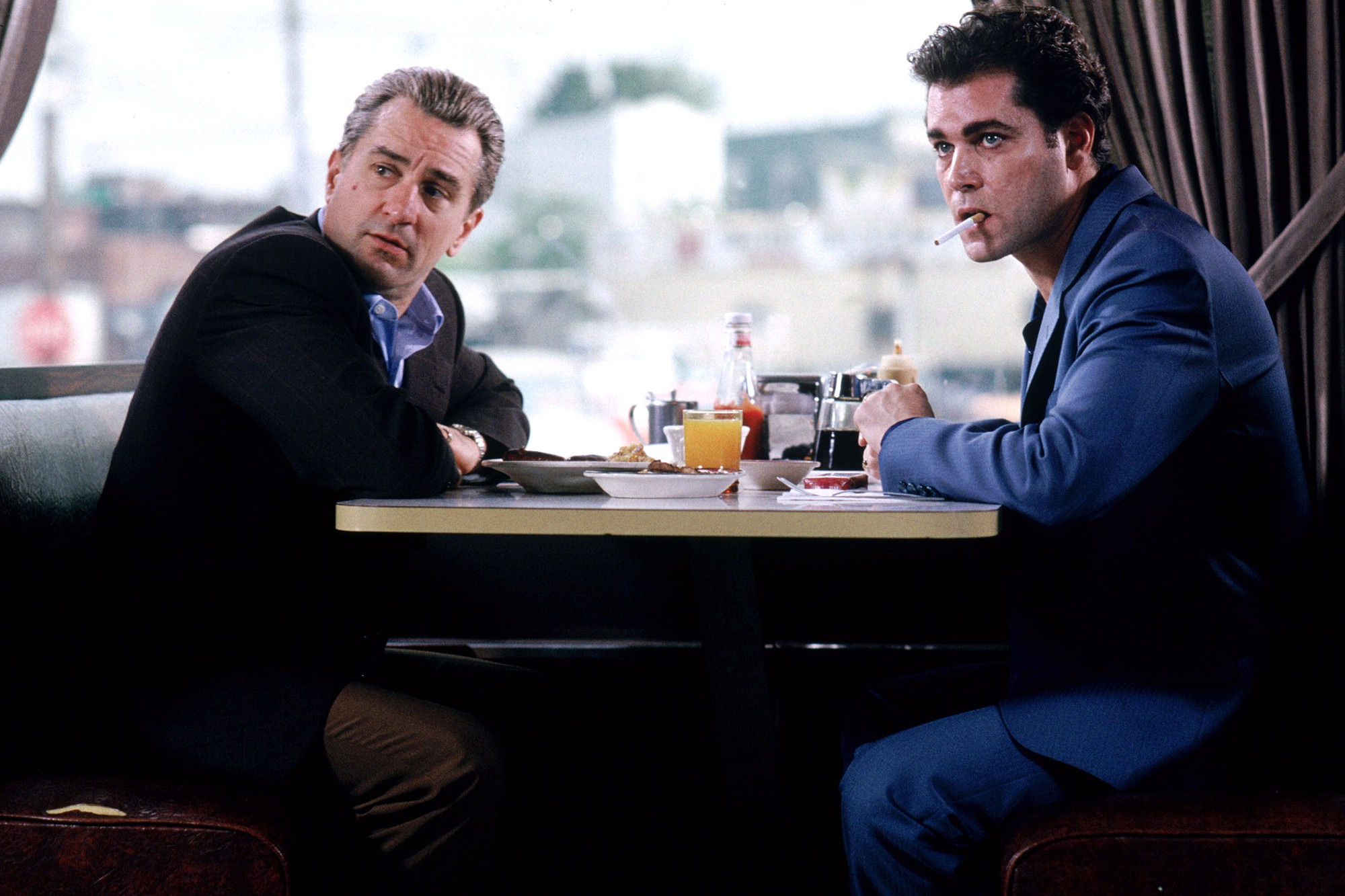 Two men sit at a diner table, one in a black suit and the other in a blue jacket, sharing an intense conversation over breakfast.