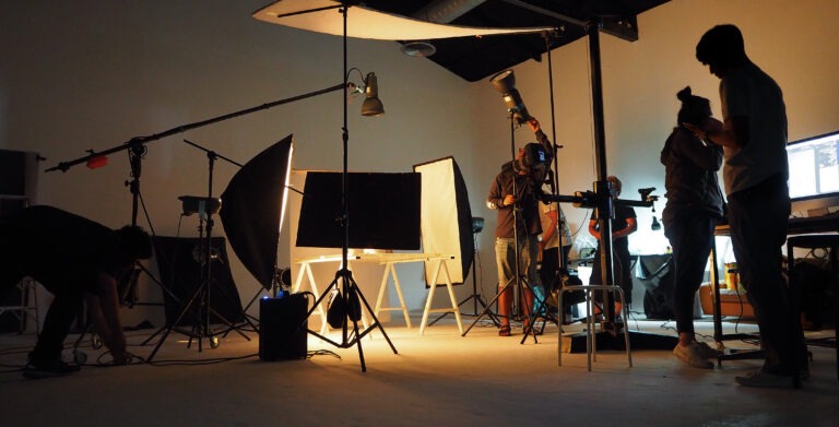 Photography studio in action, featuring multiple crew members setting up lighting and equipment for a shoot. A computer screen displays project details.