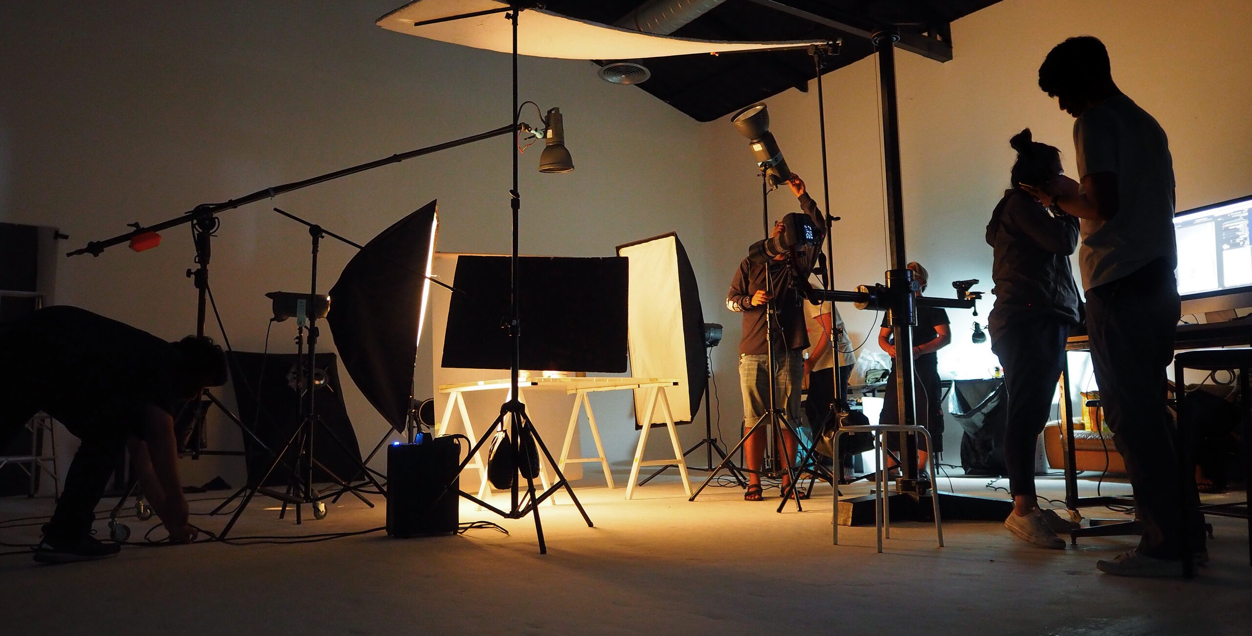 Photography studio in action, featuring multiple crew members setting up lighting and equipment for a shoot. A computer screen displays project details.